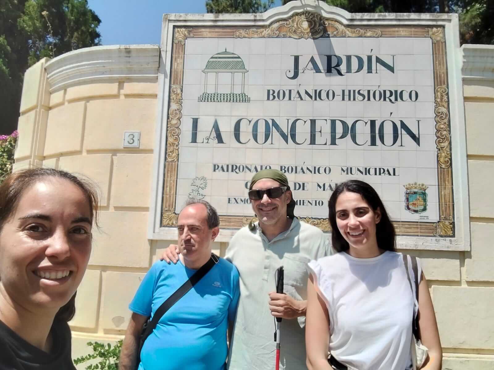 De derecha a izquierda, aparece Irene (GILSE), José María (PSC), José Miguel (PSC) y Águeda María (voluntaria) sonrientes posan frente al cartel de entrada del Jardín Botánico-Histórico La Concepción de Málaga. El cartel de azulejos blancos con letras verdes y negras está integrado en una pared clara, y se puede leer claramente el nombre del lugar. Una de las personas lleva bastón blanco, identificándose como persona con sordociega. El grupo muestra un ambiente alegre en un día soleado.
